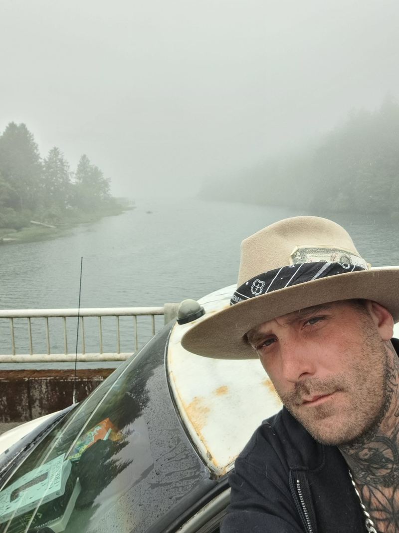 Scoop in a wide-brim hat taking a selfie beside the van on a foggy morning by a river lined with evergreens